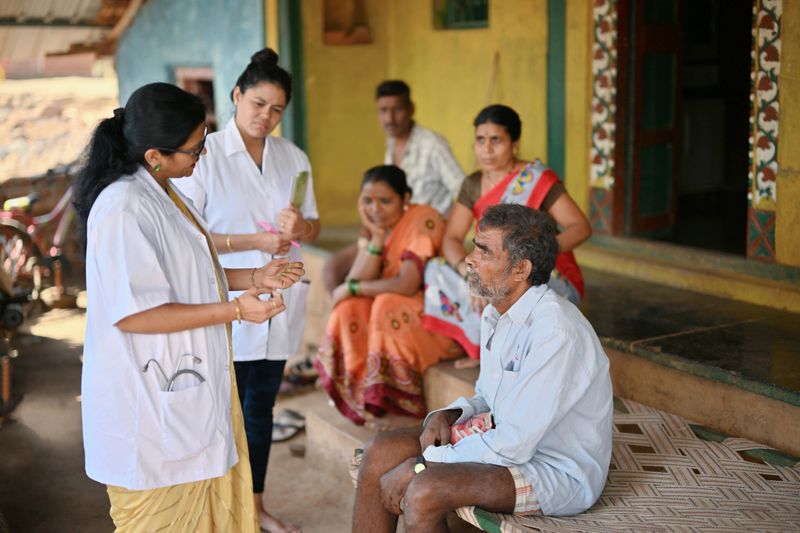 The scene conveys a sense of community engagement, with the doctors offering personalized care and health education to improve the well-being of the village people in their familiar environment
