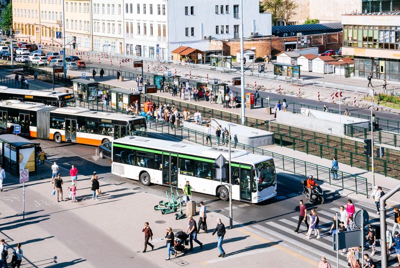 Street scene in the heart of Gdansk's city centre. The image focuses on a bustling section of the street, where multiple buses arrive and depart from the bus stop. Adjacent to it, a busy tram stop can be seen, crowded with people waiting for their trams. The pedestrian areas are filled with a diverse flow of individuals, with some crossing the road. This non-fictional depiction offers a glimpse into the energetic atmosphere of Gdansk's urban environment, highlighting the dynamic transportation system and the lively street activity in the city centre.