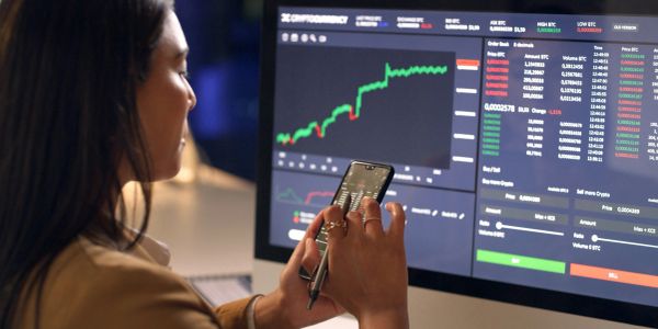 Woman analyzing cryptocurrency market charts on phone and computer.