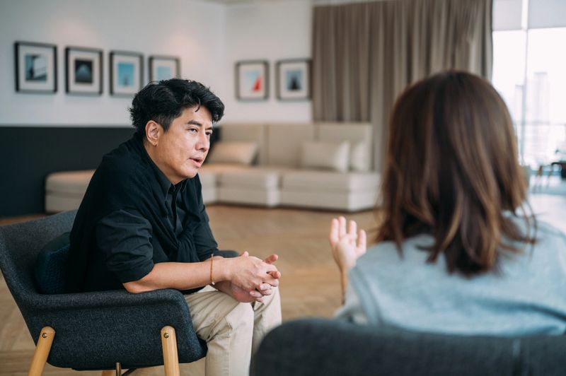 An Asian man sitting on cozy sofa and discussing faced problem with highly professional psychologist during therapy session at cozy office.