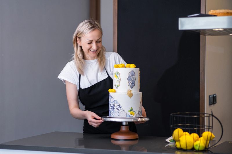 Happy female confectioner looking at finished wedding cake decorated with yellow lemons blonde woman smiling standing at kitchen table and enjoying result of long work