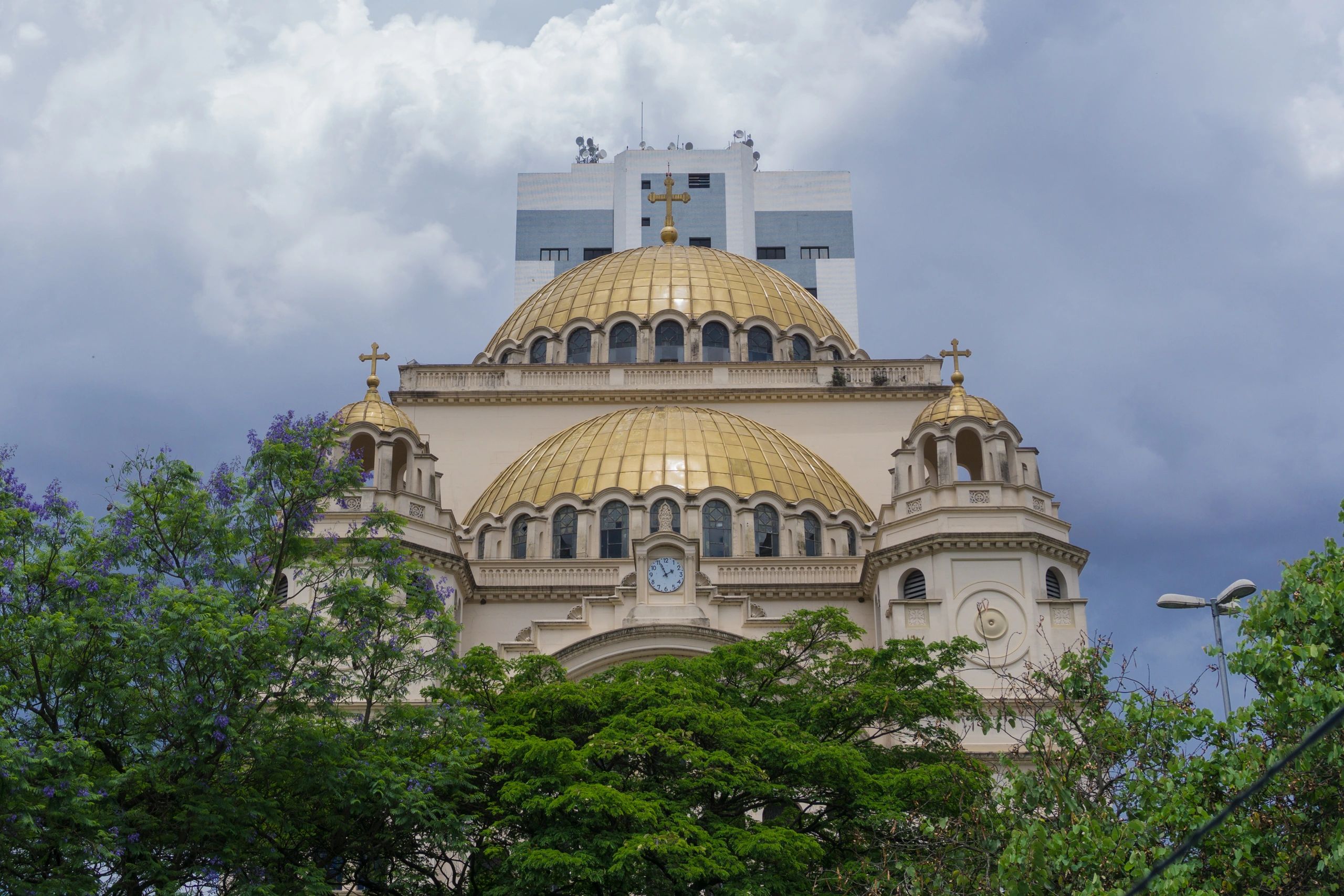 Golden-domed church with crosses and a clock, framed by green trees against a cloudy sky.