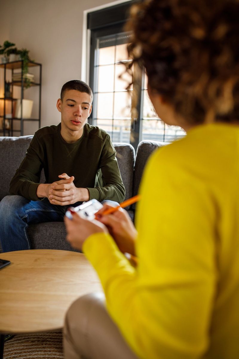 Over the shoulder view of mid adult female therapist giving constructive advice to insecure teenage boy, that is struggling with negative emotions, sitting across from her on the sofa, during a therapy session.