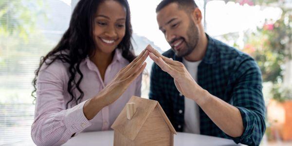 Couple protecting a miniature wooden house with their hands.