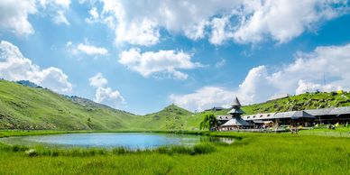 A serene lake surrounded by lush green hills and a quaint building under a bright blue sky.