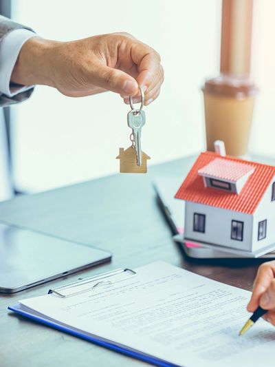 Person handing over house keys while another signs a document with a house model nearby.
