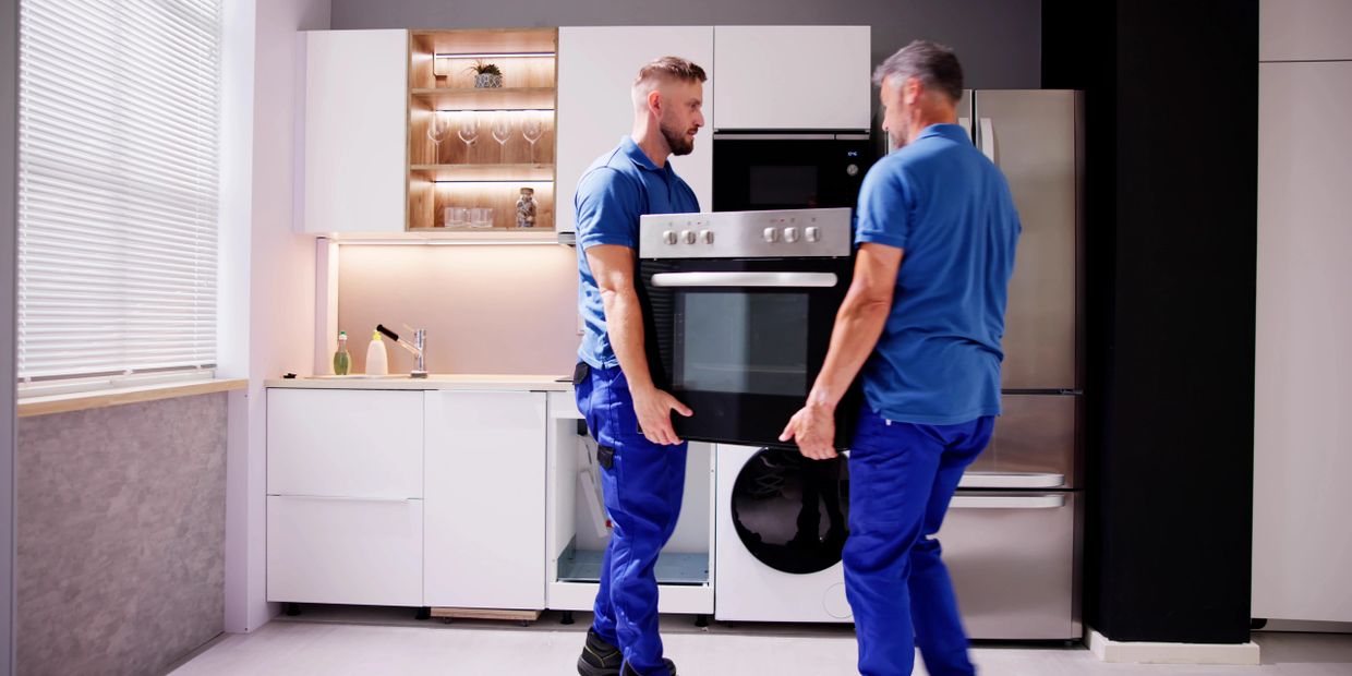 Two workers in blue uniforms carrying an oven in a modern kitchen.