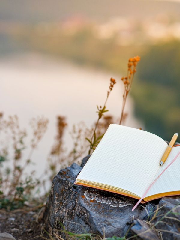 Open notebook with a pen on rocks overlooking a river and blurred nature.