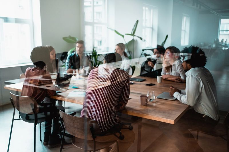 Group of young business people working together in creative office. Multi-ethnic group of business persons on a meeting in modern office. Shot of a group of people sitting on a conference table in board room. The view is through glass.
