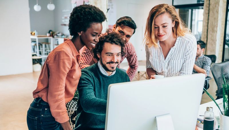 Shot of a group of colleagues using computer together at work. Four coworkers working on a computer in the office. Multi-ethnic creative team using computer in modern office. Young smiling business people cooperating while working on PC.