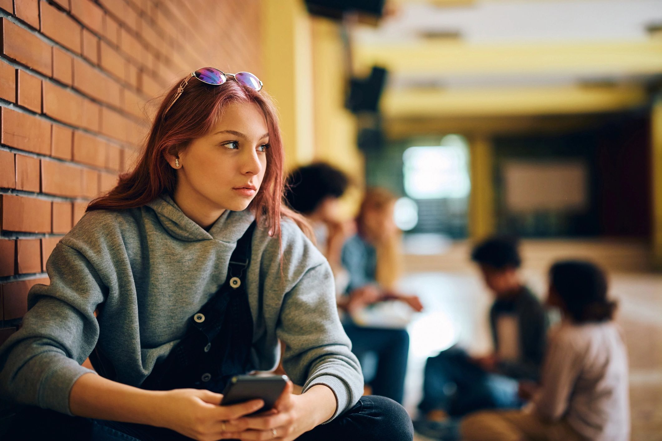 Teen girl with phone sitting against brick wall, looking away thoughtfully.