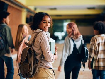 Smiling student with backpack holding books, walking with friends in school hallway.