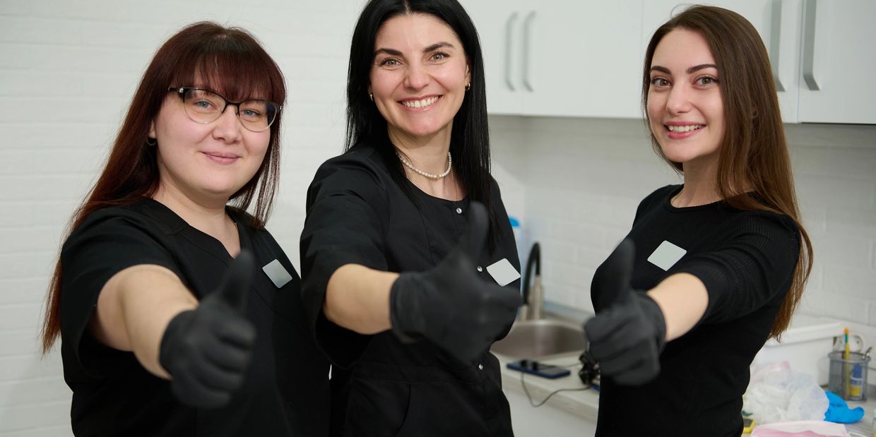 Three smiling women in black gloves giving thumbs up in a clinical setting.
