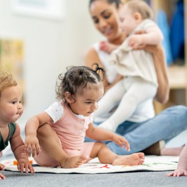 Babies sitting and playing on the floor with a woman holding a baby.