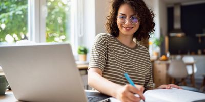 Young woman with glasses smiling while working on a laptop and writing in a notebook.