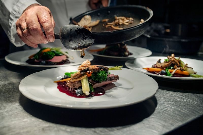 Chef at the pass preparing a dish of venison served with mushrooms, broccoli, carrots and new potatoes with a seasonal berry sauce and decorated with fresh pea shoots. Colour, horizontal format with some copy space. Photographed on location at a restaurant on the island of Moen in Denmark.