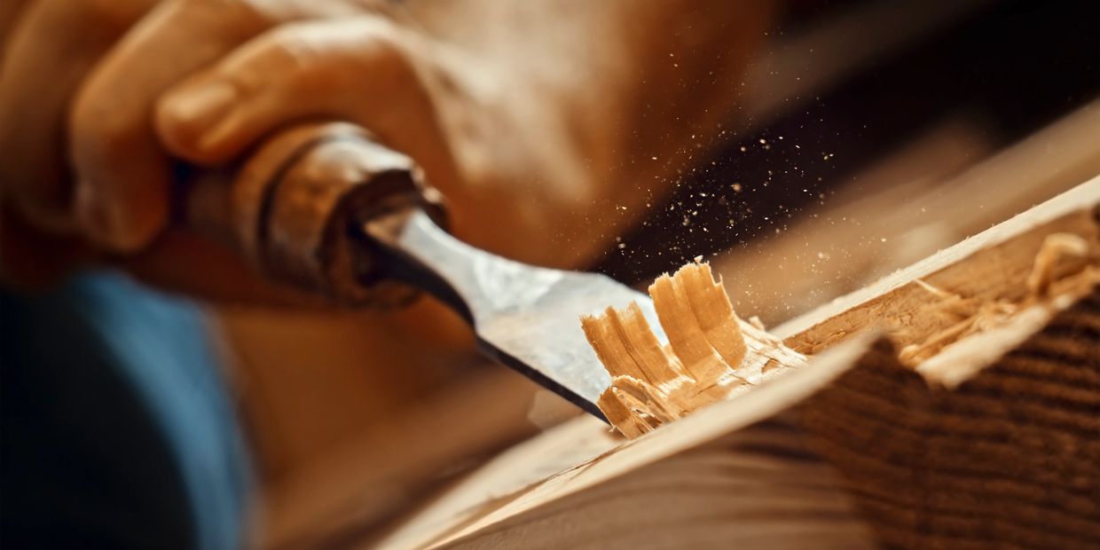 Close-up of a hand carving wood with a chisel, producing fine wood shavings.