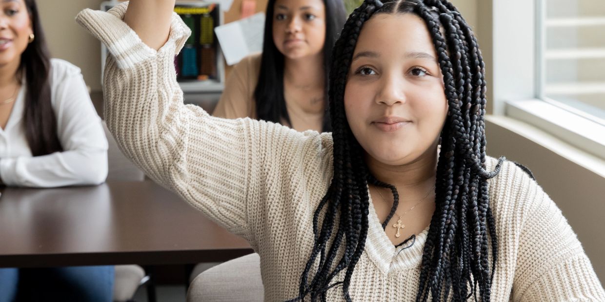 A young woman with braided hair raises her hand attentively in a classroom setting.