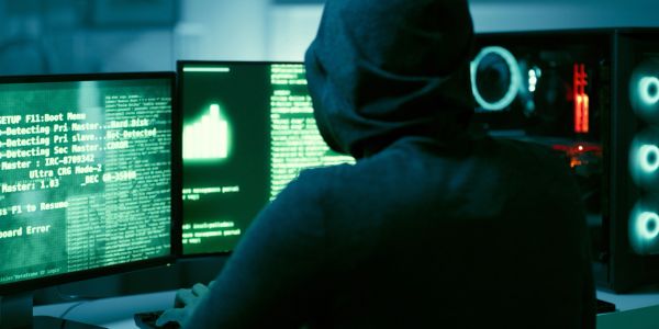 Hacker in a hoodie working on multiple computer screens.