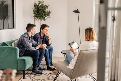 Two men in therapy talking to a therapist holding a tablet in a cozy office.