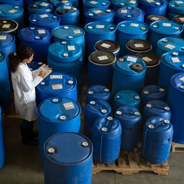 Woman in lab coat checks labels on numerous blue barrels in a warehouse.