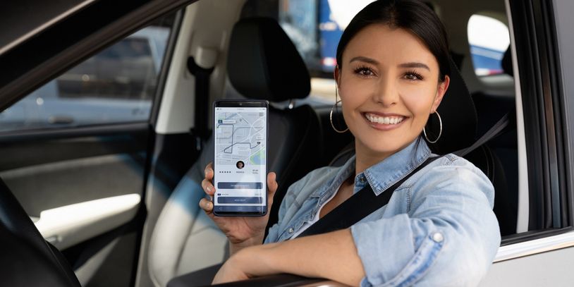 Mujer sonriente en coche mostrando una app en su celular.