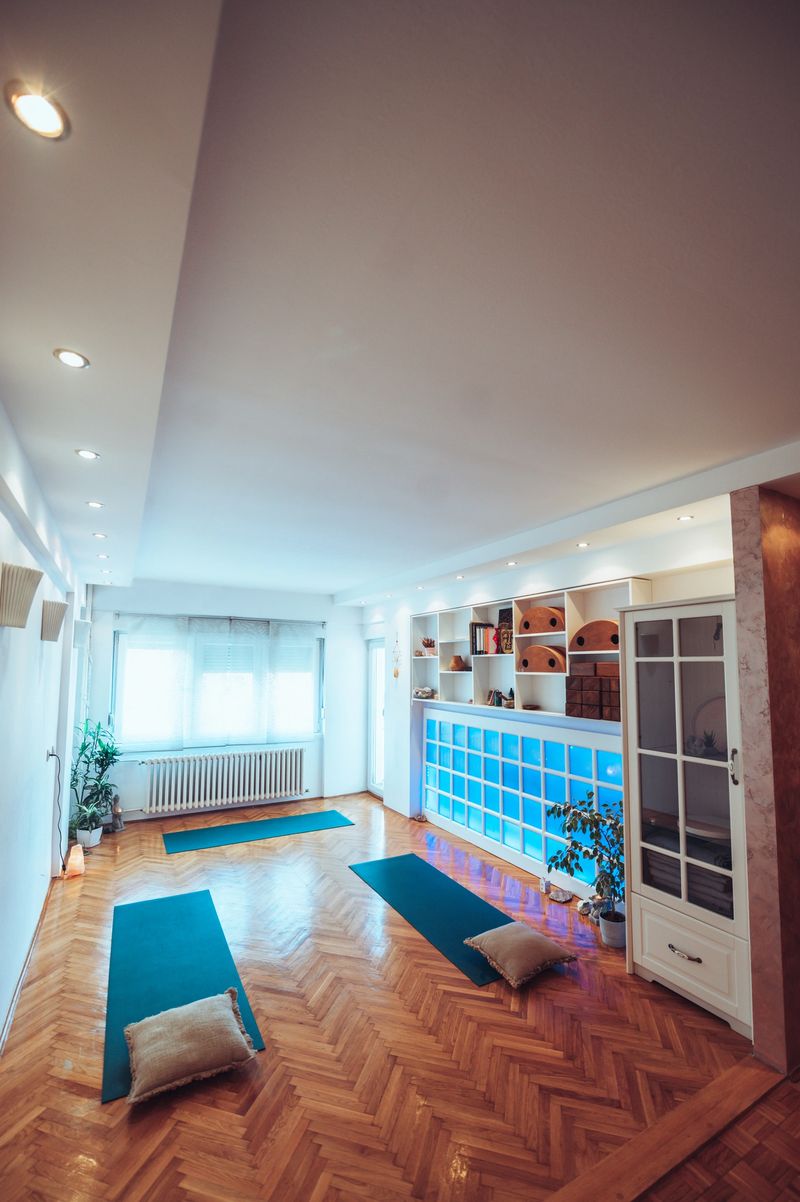 Wide shot of a cozy and bright yoga studio space with big window and wooden floor. Long white shelf on the right wall with yoga equipment and books. Blue yoga mats on the floor and plants.Copy space.