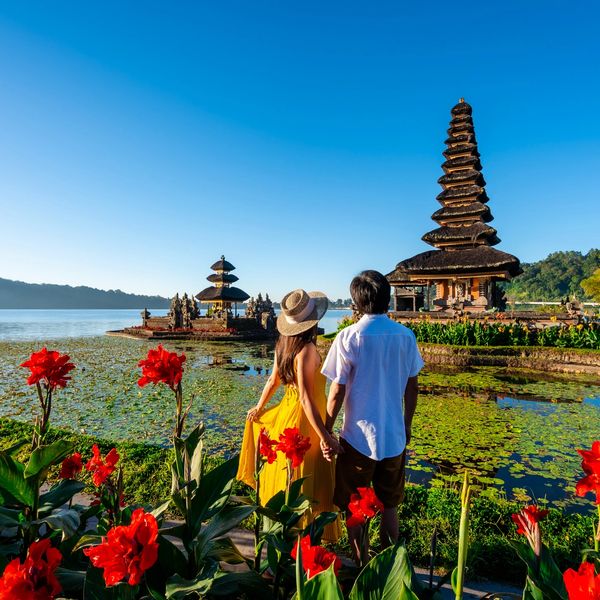 Couple holding hands near water temple with red flowers and clear blue sky.