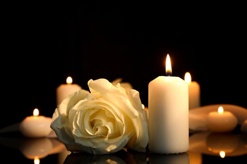 White rose and burning candles on black mirror surface in darkness, closeup with space for text. Funeral symbols