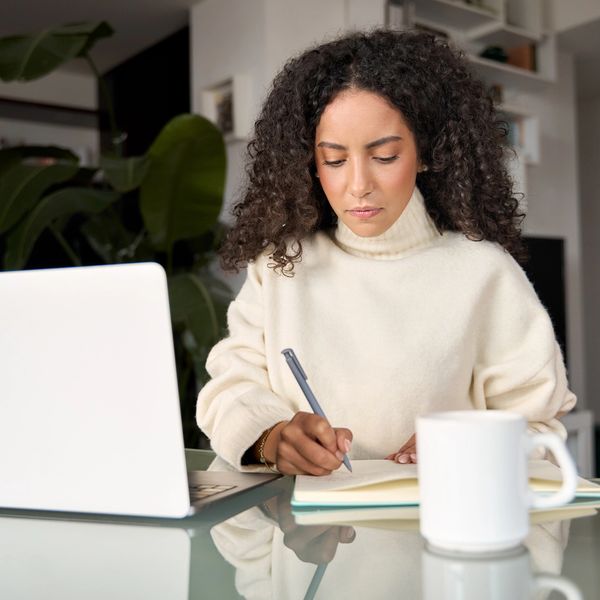 Woman sitting at a table with her laptop, a notebook, and a cup of coffee. She is taking notes.