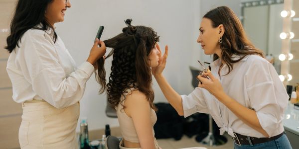Two stylists working on a woman's hair and makeup.