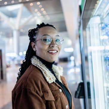 Smiling woman with glasses and braided hair stands near a refrigerated display of water bottles.