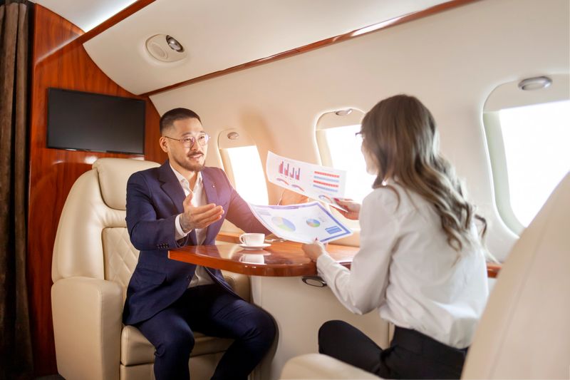 business meeting in airplane, asian businessman in private jet is talking with female colleague and looking through charts and documents, business people flying in airplane and working with papers