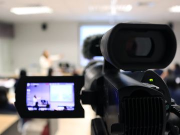Video camera recording a person giving a presentation in a classroom.