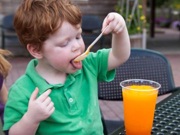 Young boy in green shirt eating orange sherbet outdoors.