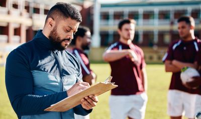 Coach writing on clipboard while players stand in background on sports field.