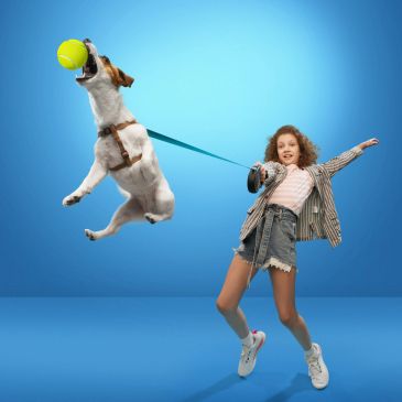 Energetic dog leaps to catch a ball while girl holds the leash with excitement.