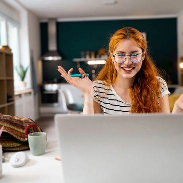 A young woman with red hair smiling and gesturing during a video call at home.