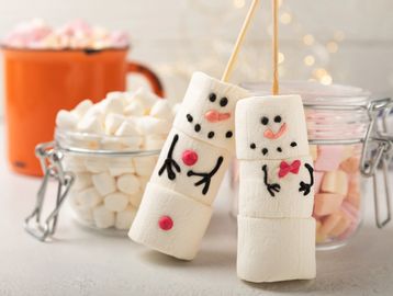 Marshmallow snowmen on sticks with jars of mini marshmallows and a mug in the background.