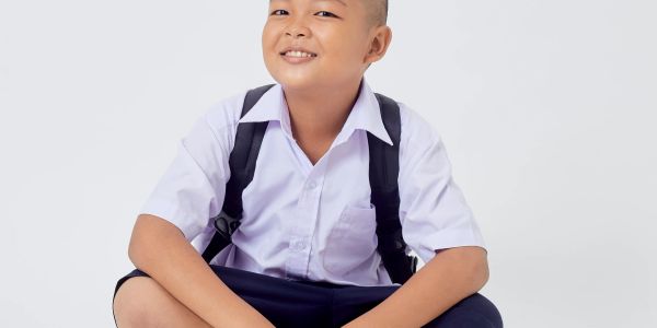 Smiling schoolboy in uniform sitting cross-legged with backpack on white background.
