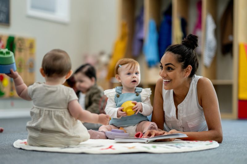 A small group of babies sit on the floor playing with various toys while their teacher supervises and engages them.  They are each dressed casually  and appear content.