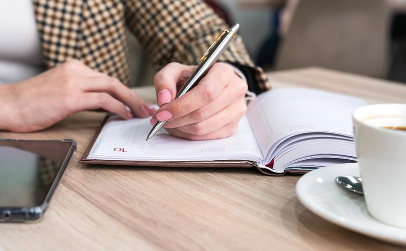 A young left-handed girl writes a plan for the day in a notebook while sitting in a cafe. Close-up. Selective focus.