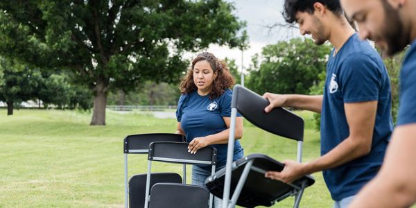 Three people setting up black chairs outdoors on a grassy field.
