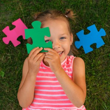 Smiling girl holding green puzzle piece, lying on grass with colorful puzzle pieces around.