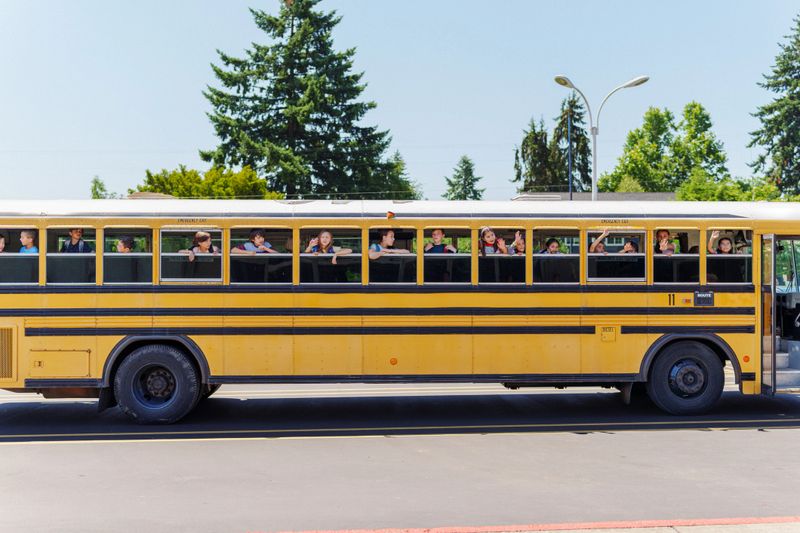 A multiracial group of elementary age boys and girls smile and wave out open windows of a parked yellow school bus on a sunny spring day in the Pacific Northwest region of the United States.