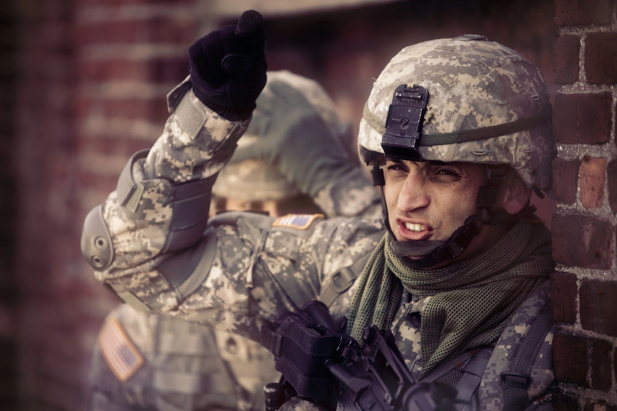 A soldier signaling with his hand near a brick wall.