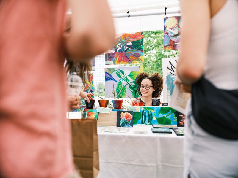 Young Latin woman artist selling her art at Outdoor Market.  She is dressed in casual East Indian dress. Exterior of public park in downtown Toronto, Canada.