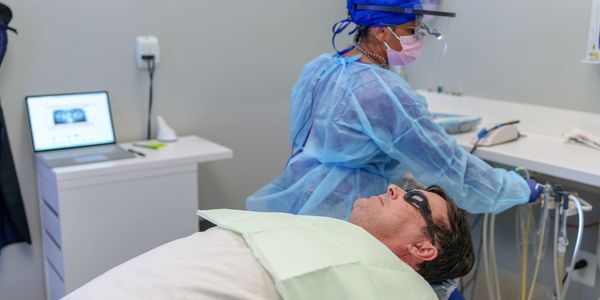 A man wearing protective eyewear lies in a dental chair while a dentist in PPE prepares equipment.