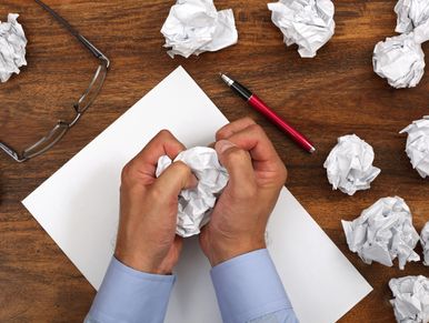 Frustrated person crumpling paper amid discarded drafts on desk.