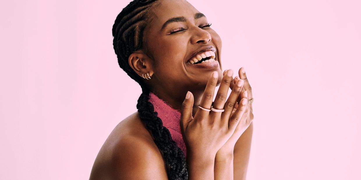 Joyful woman laughing with hands clasped against a pink background.